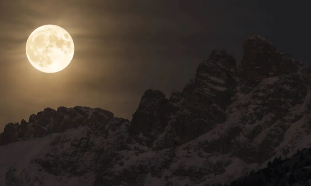 Full Moon glowing above snowy mountains, reflecting sunlight in the night sky