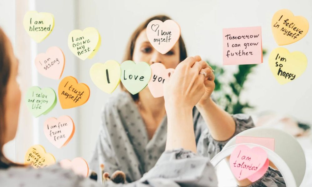 Woman placing heart-shaped sticky notes with positive affirmations like "I love you" and "I am strong" on a mirror, symbolizing mindful use of Words of Affirmation